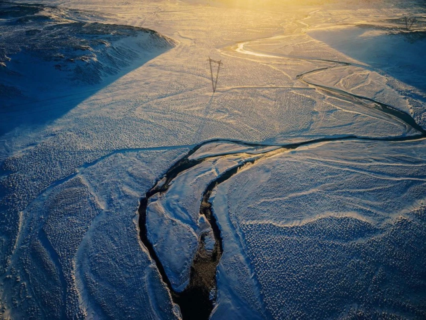Aerial view of snowy landscape with mountains, a frozen river, and a transmission tower under bright sunlight, creating long shadows.