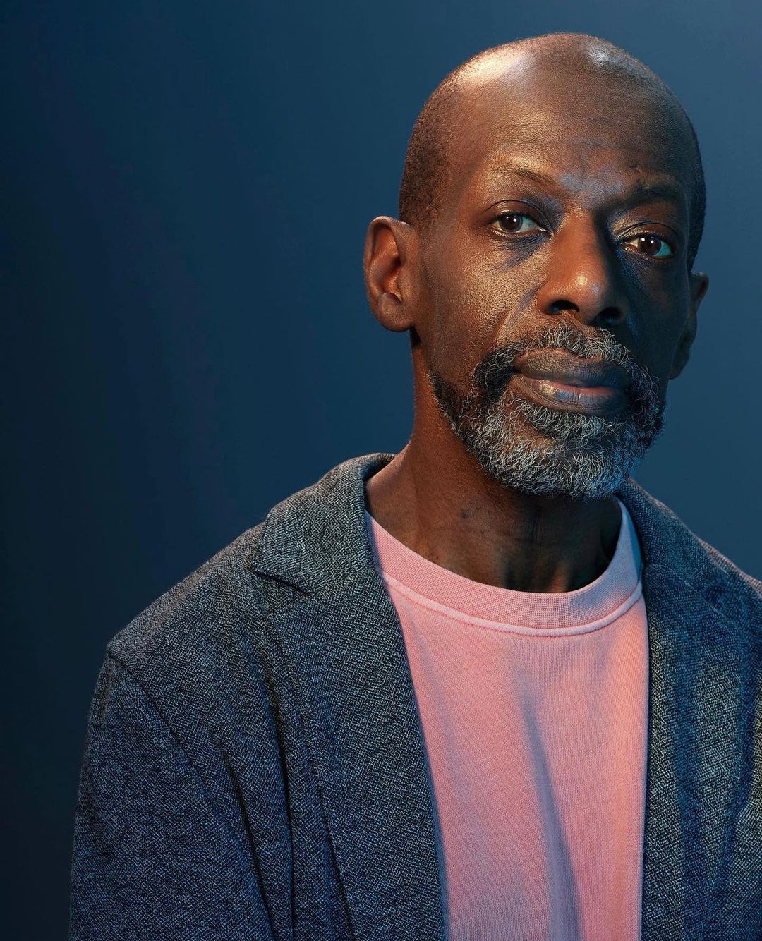 Portrait of a mature adult African-American man with salt and pepper beard wearing a grey blazer and light pink shirt against a dark blue backdrop.