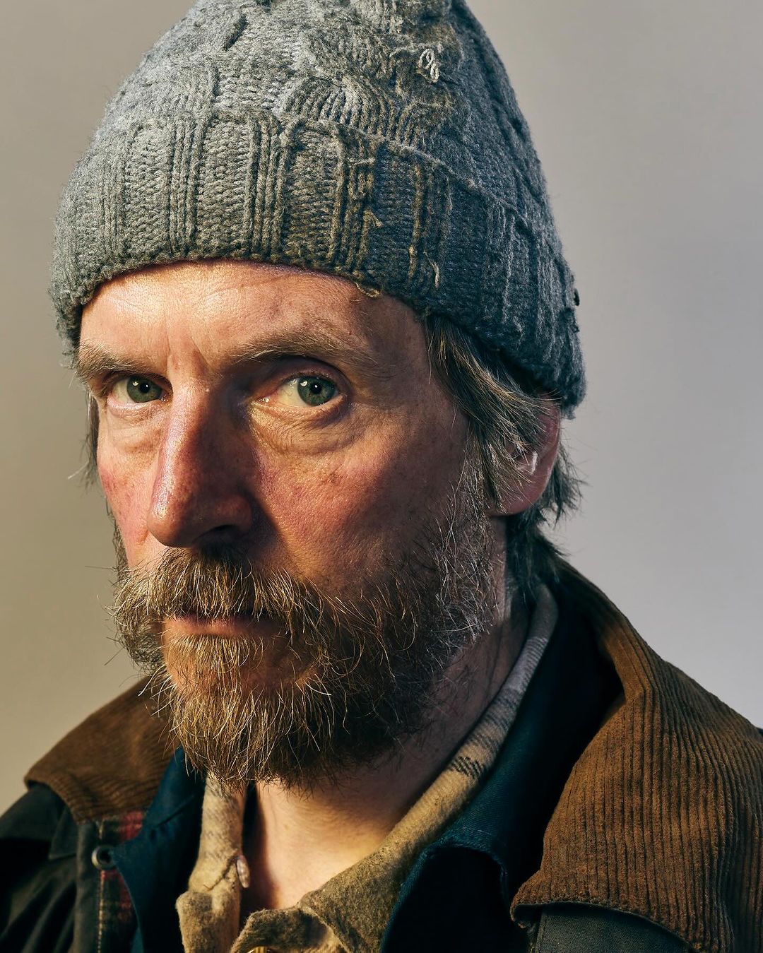 Portrait of an adult man with a beard wearing a gray knit hat and a corduroy jacket, captured in a close-up shot with soft, diffused lighting.