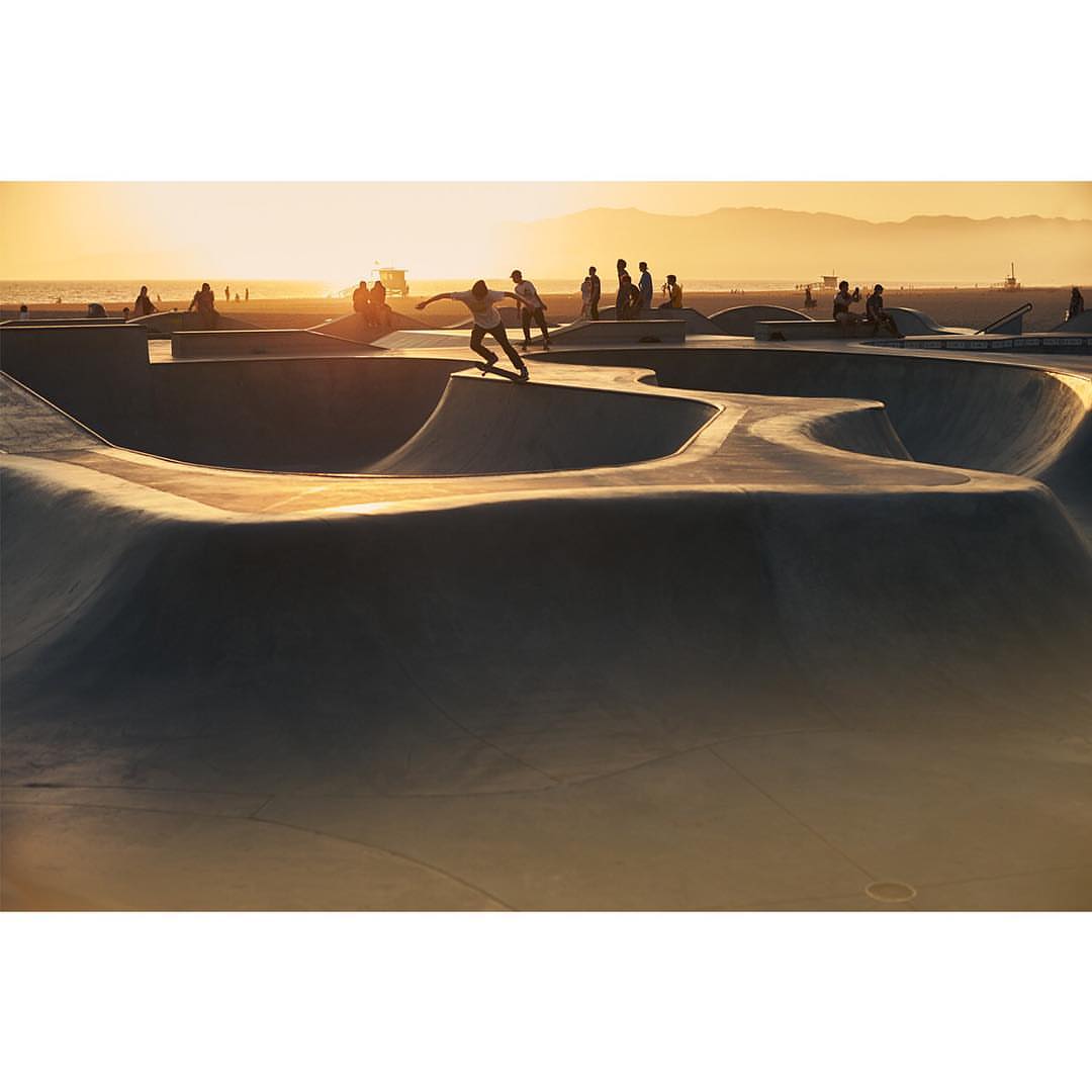 A skateboarder performs a trick at a skate park during sunset. The park is crowded with people, and the mountains in the background.
