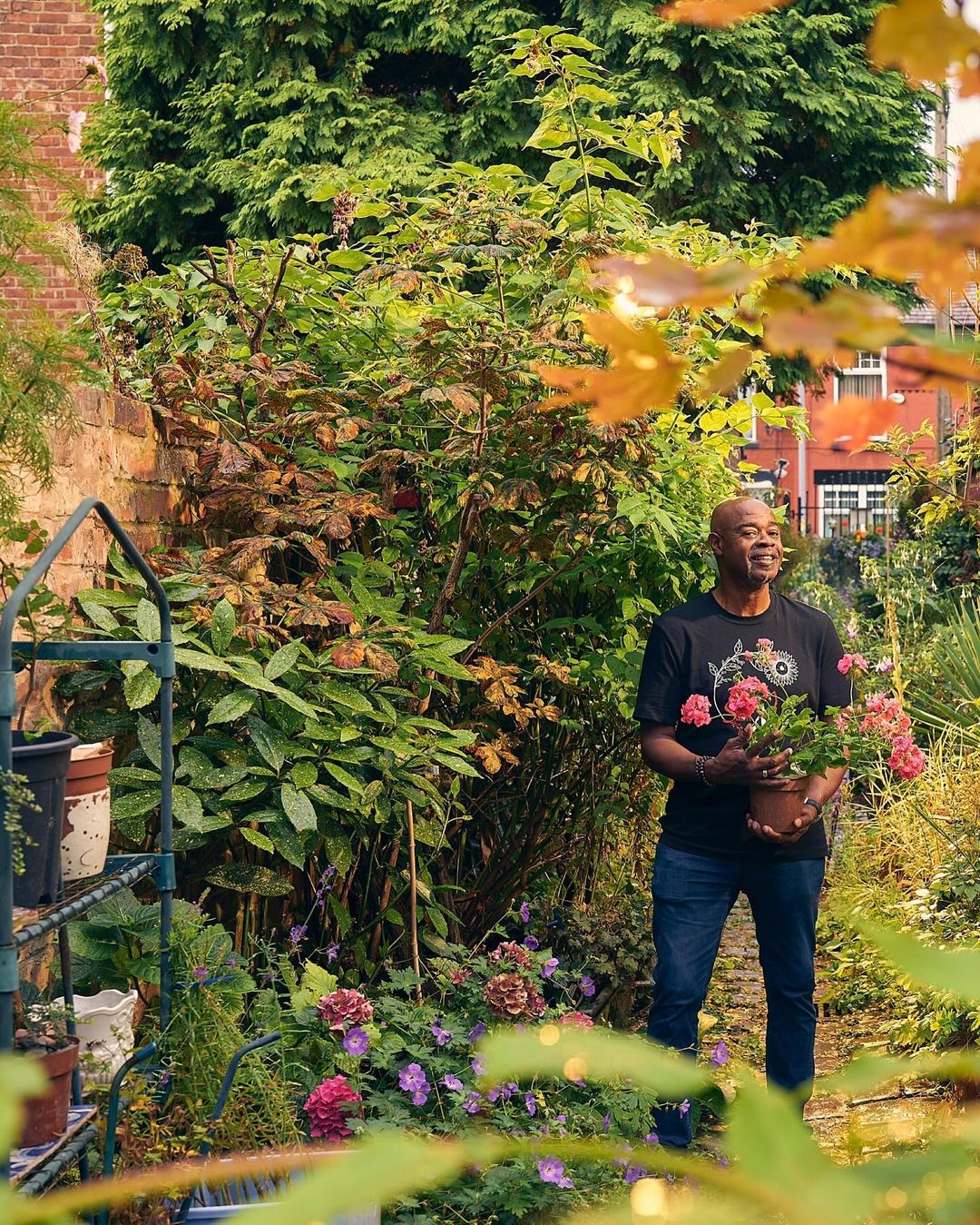 Middle-aged Black man in a garden holding a potted plant with roses, surrounded by lush greenery and foliage on a sunny day.