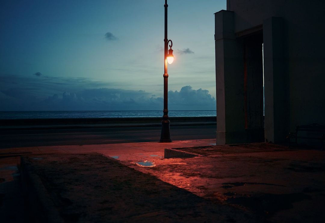 Nighttime scene by the ocean featuring a vintage streetlamp casting a warm glow on an empty street with a building in the background.