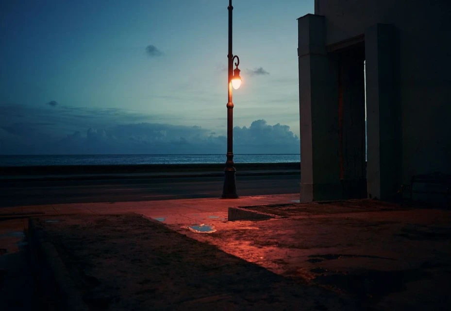 Nighttime scene by the ocean featuring a vintage streetlamp casting a warm glow on an empty street with a building in the background.