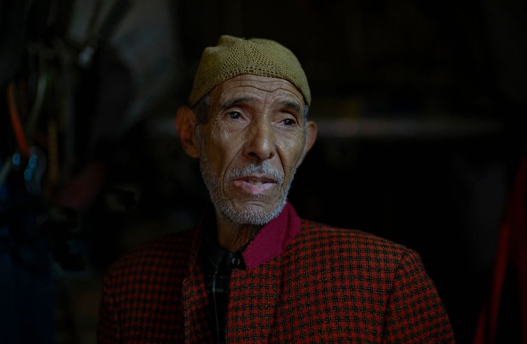 Portrait of an elderly man with weathered skin wearing a patterned jacket and knit hat against a dark background, close-up shot.