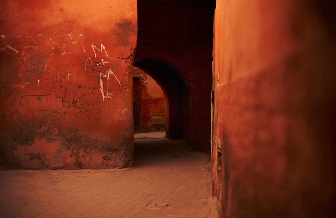 An atmospheric view down a narrow alleyway with arched openings and textured orange walls featuring painted white graffiti.
