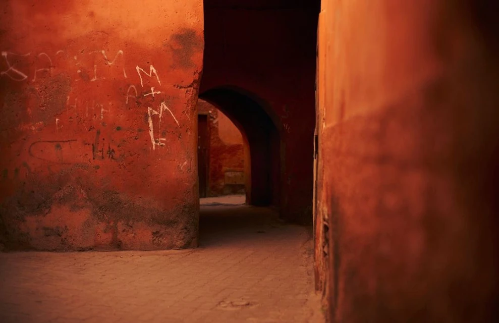 An atmospheric view down a narrow alleyway with arched openings and textured orange walls featuring painted white graffiti.