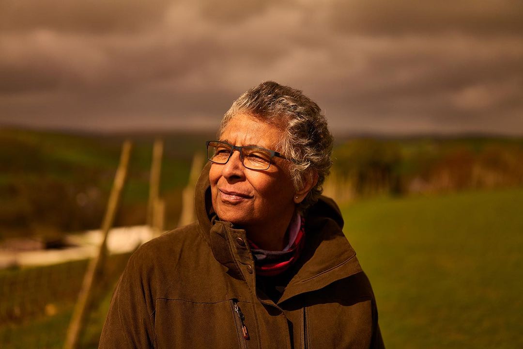 Outdoor portrait of a mature adult woman wearing eyeglasses and a brown jacket, smiling slightly, set against a rural landscape background.