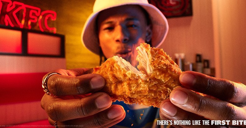 Close-up of a person holding a fried chicken piece, partially bitten, with a KFC sign in the background, food presentation.