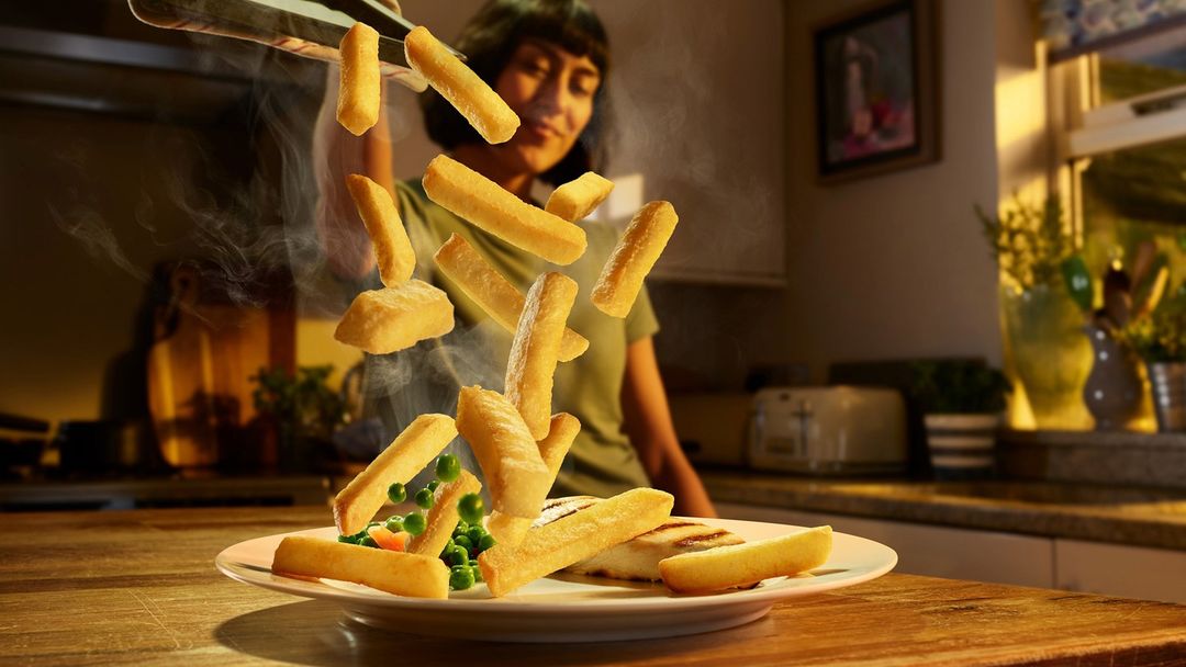 Woman in kitchen tosses chips into a plate with peas and fish. Light illuminates the food, creating a warm, inviting scene.