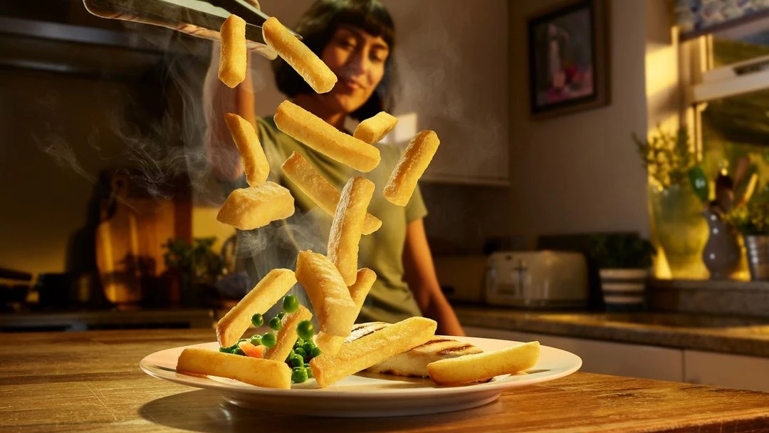 Woman in kitchen tosses chips into a plate with peas and fish. Light illuminates the food, creating a warm, inviting scene.