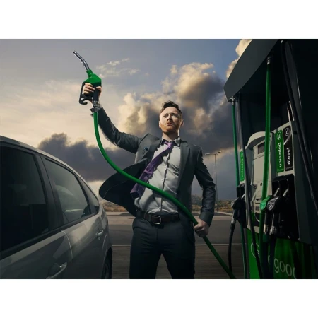 Man in suit triumphantly holds up gas pump nozzle at a gas station on a cloudy day near a gray car, symbolizing success or satire.