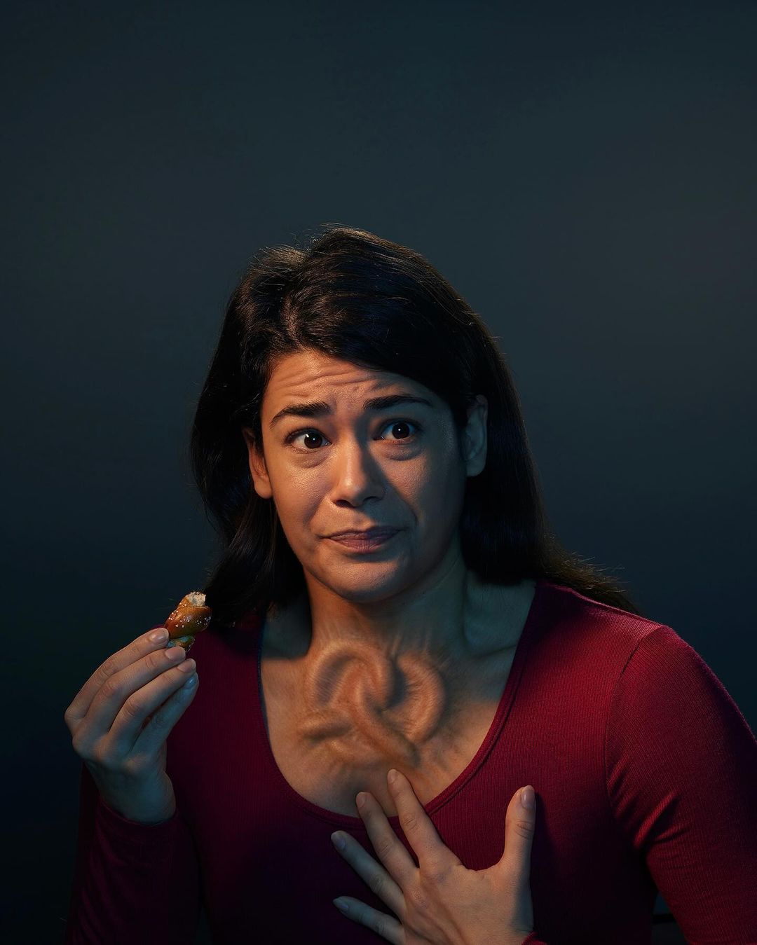 Adult woman with pained facial expression holds food with pretzel shaped deformity in her chest area against dark background.
