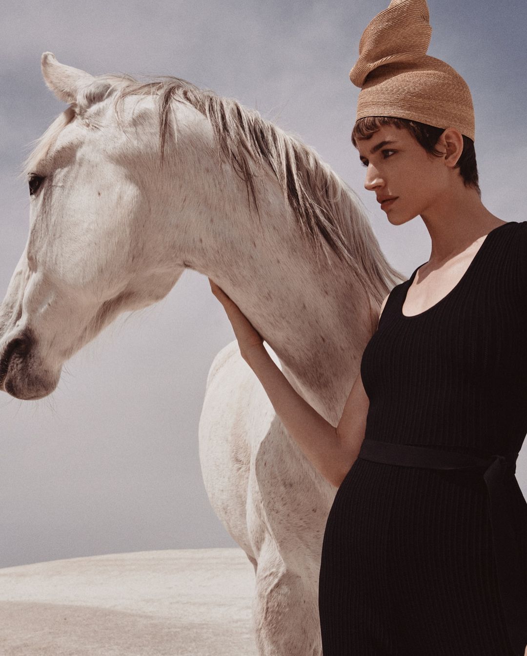 A woman in a black dress stands next to a white horse in an outdoor setting. The scene evokes a sense of fashion and equine elegance.