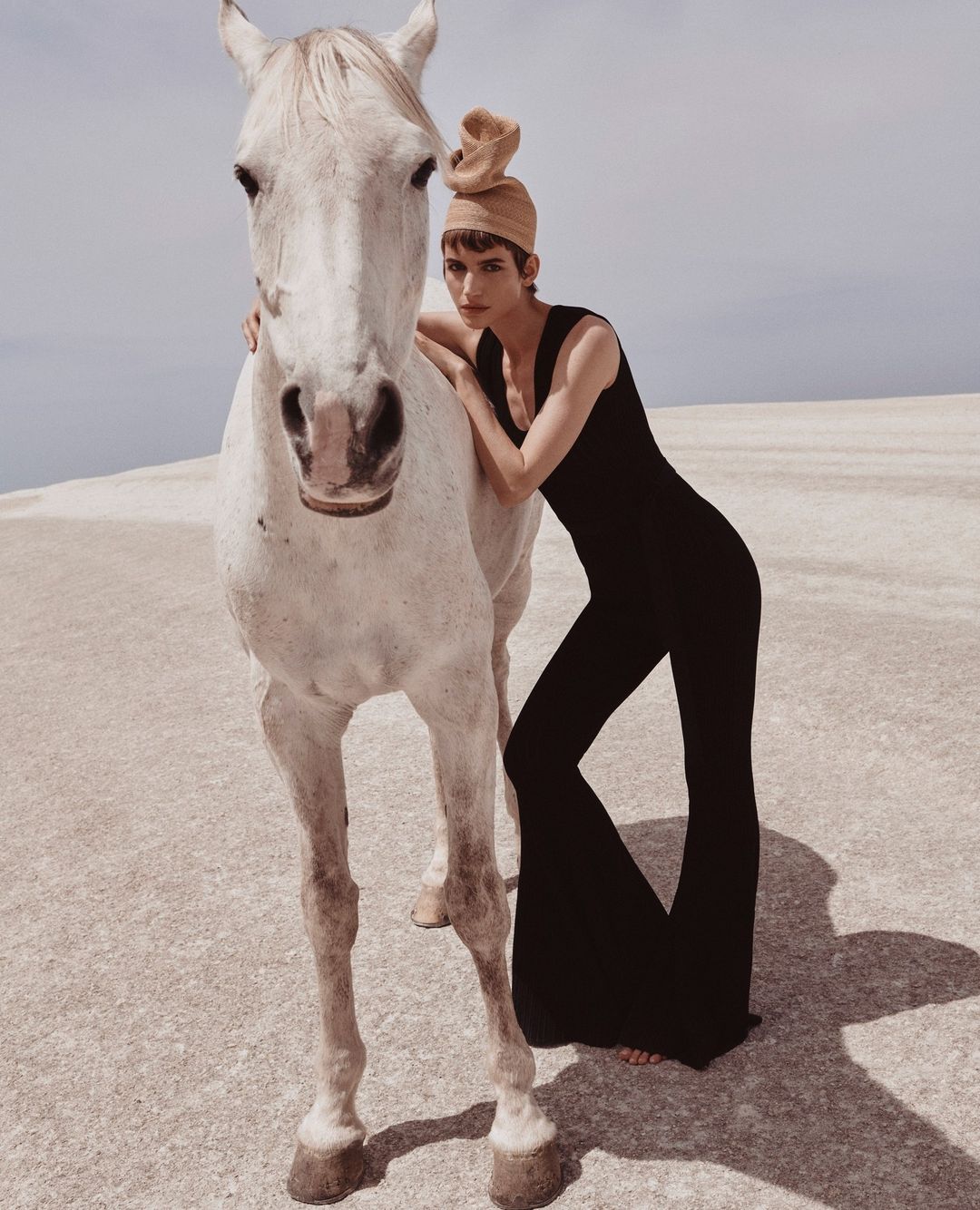 Fashion model in black jumpsuit poses with a white horse against a neutral horizon in an outdoor fashion editorial shot.