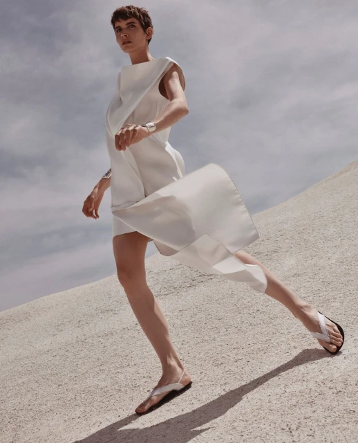 A fashion shot showcases a woman in a flowing white dress and sandals walking on a sandy surface against a backdrop of a cloudy sky.