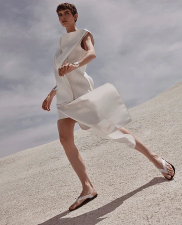 Model in a white sleeveless dress stepping forward on a sand dune under an overcast sky by Alex Waltl