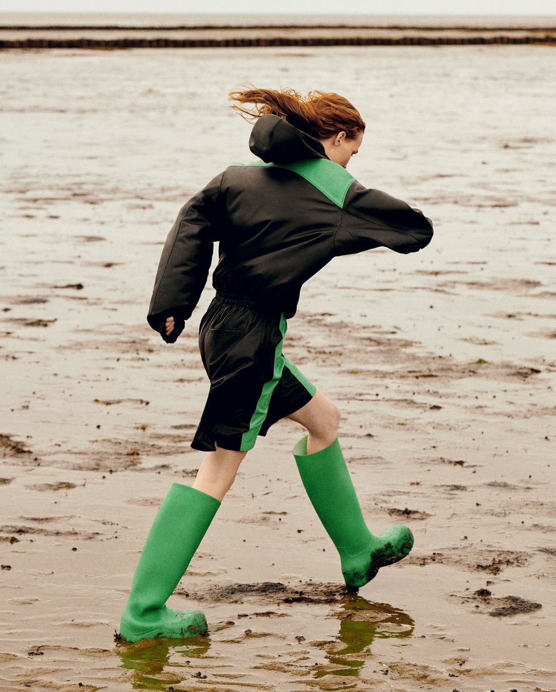 Woman in black and green jacket and green boots running across a sandy, muddy beach, with neutral sky and distant horizon.
