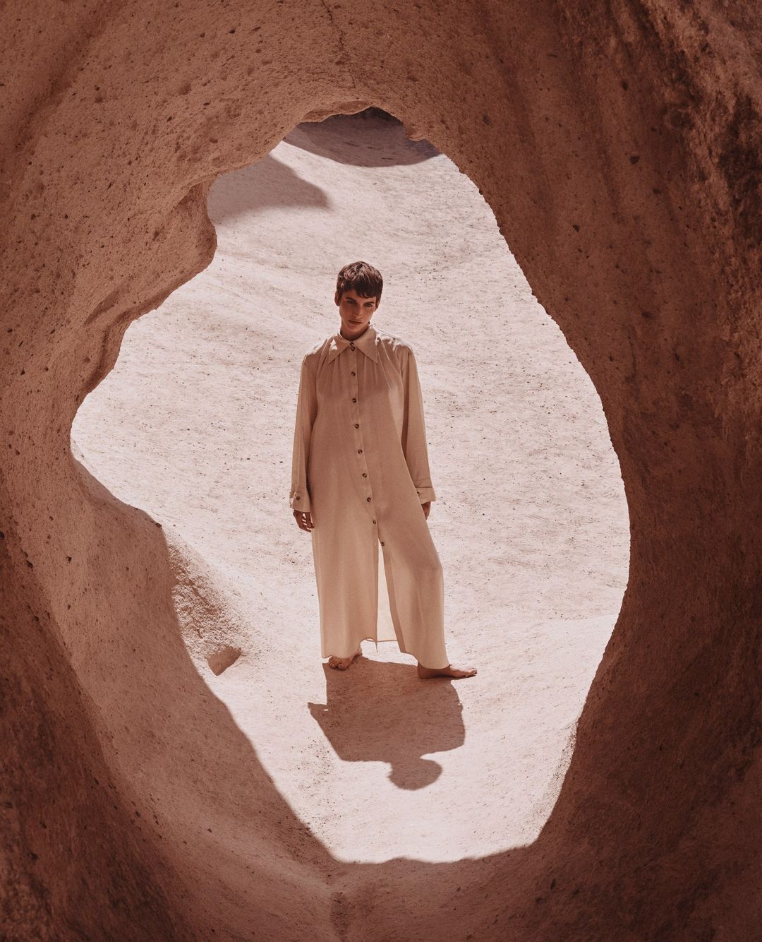 Fashion shot of a woman in a tan jumpsuit standing in a cave with neutral rock formations, captured in a medium shot.