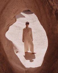 Model wearing long cream button-up tunic standing inside a natural rock cave opening by Alex Waltl