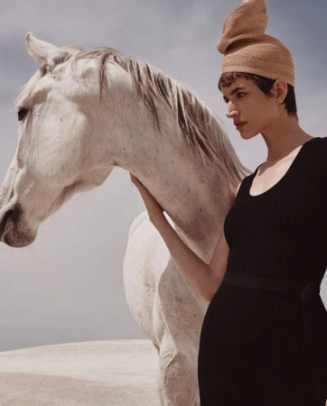 Woman in black dress and structured beige hat touching a white horse outdoors.