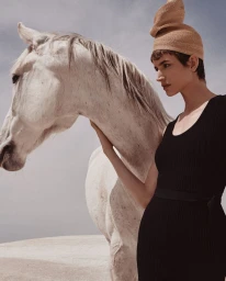 Woman in black dress and structured beige hat touching a white horse outdoors.