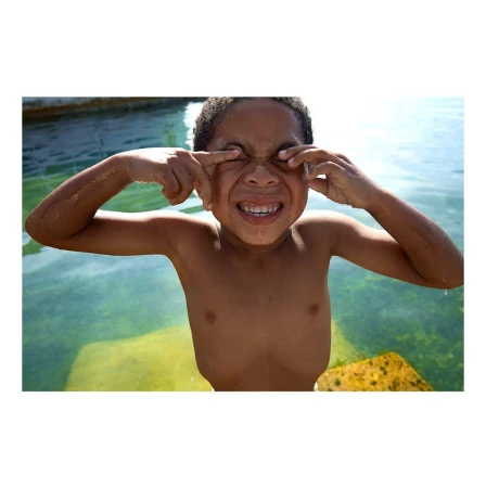 Close-up of an African-American child in waist-deep water, squinting with fingers pulling eyelids down, and smiling broadly, outdoors.