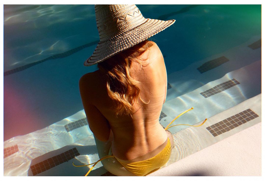 Blonde woman in yellow bikini and sun hat sits on the edge of a bright blue swimming pool enjoying the warm sunlight. 