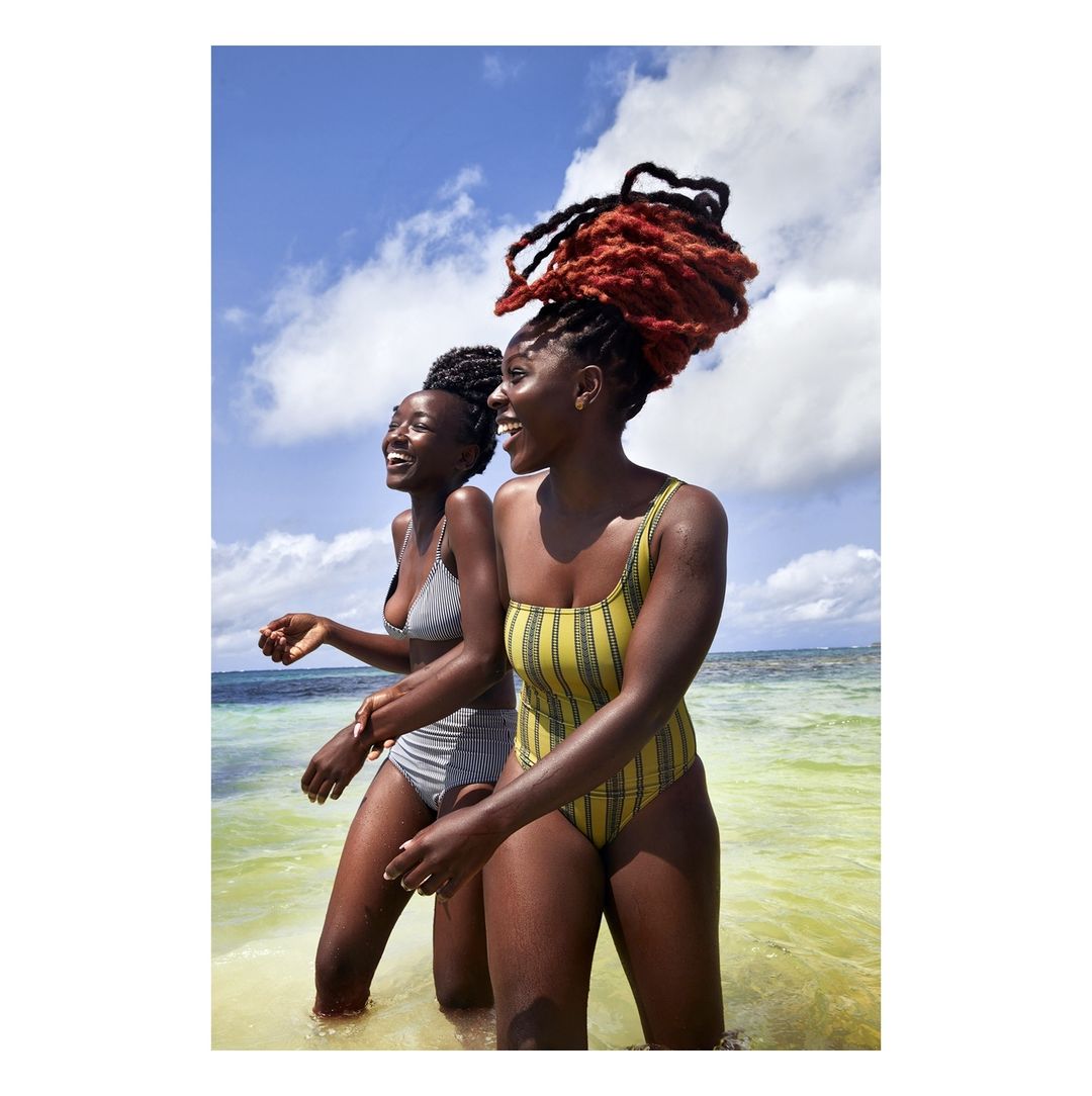 Two happy African-descent women in swimsuits stand in clear, shallow ocean water under a sunny sky with scattered clouds.