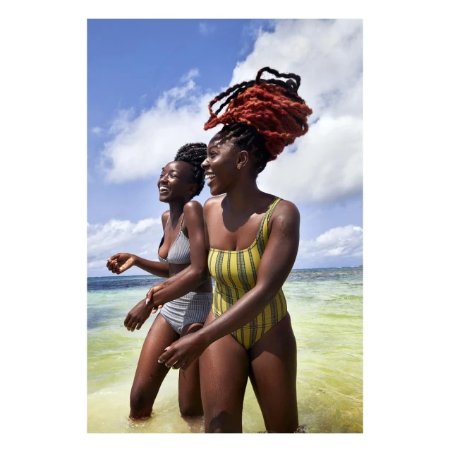 Two happy African-descent women in swimsuits stand in clear, shallow ocean water under a sunny sky with scattered clouds.