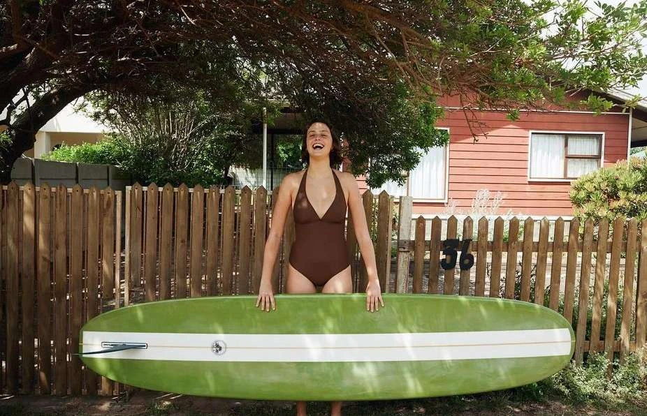A smiling woman in a brown swimsuit stands outdoors with a surfboard in front of a wooden fence and a red house.