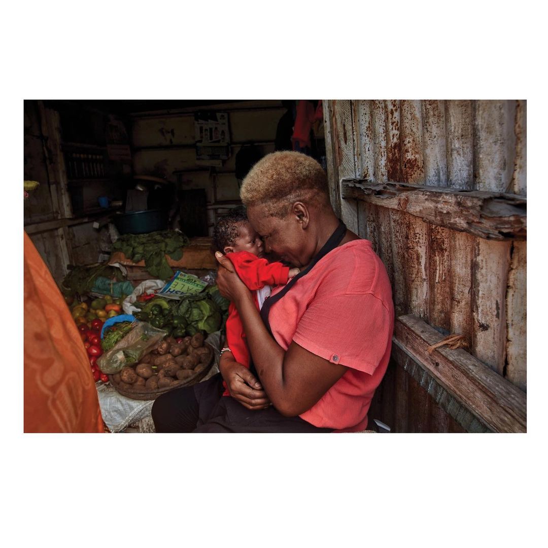 A woman holds a baby inside an open market stall. The woman wears a salmon shirt, and the baby wears red. Produce is in the background.