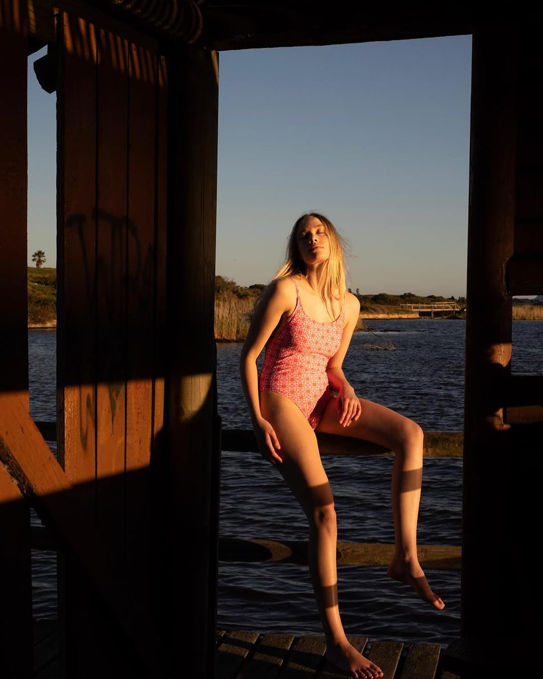 Young woman in a pink floral swimsuit sits on a wooden railing near a lake during sunset, framed by wooden pillars and shadows.