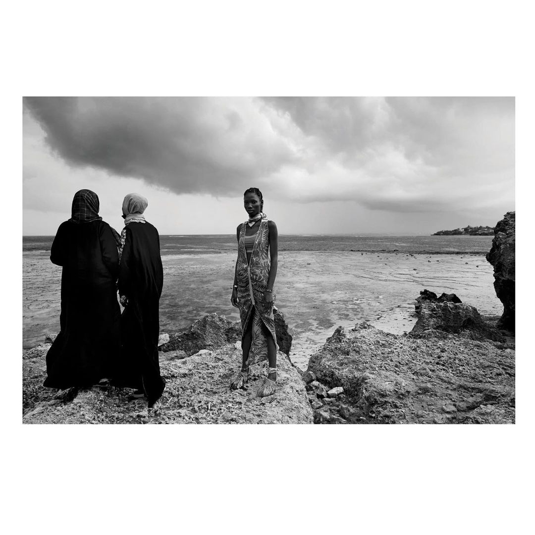 Three women on a rocky beach with cloudy sky, in black and white. One stands facing forward, two are veiled, on the ocean's edge.