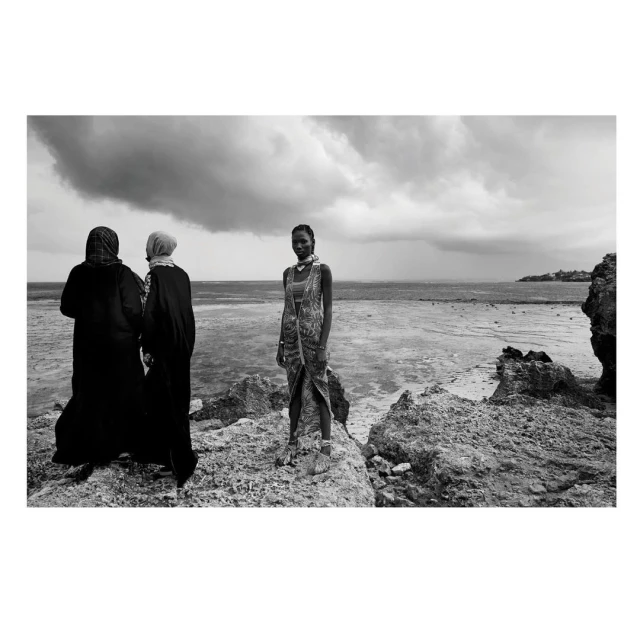 Three women on a rocky beach with cloudy sky, in black and white. One stands facing forward, two are veiled, on the ocean's edge.