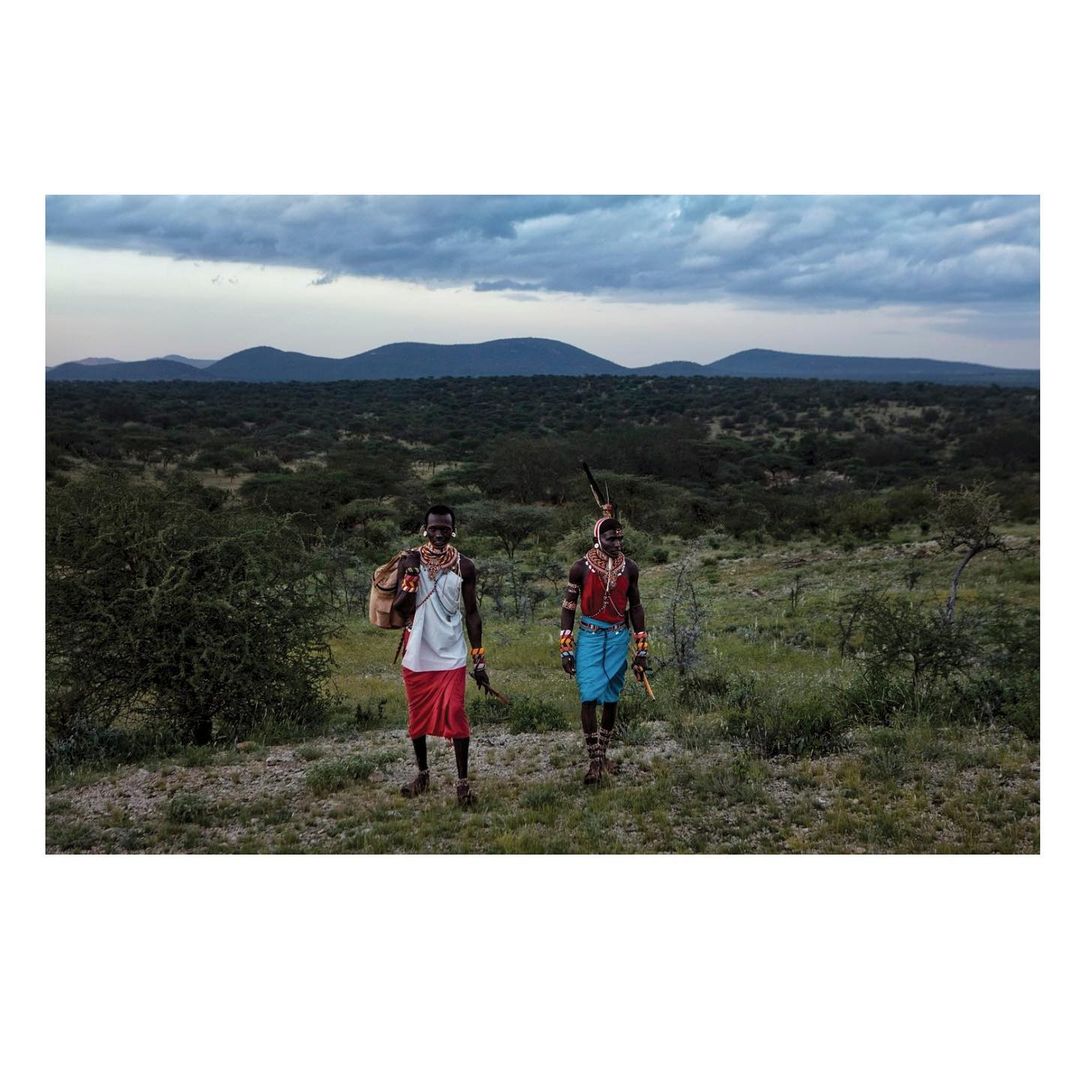 Two Samburu people in traditional dress stand in a grassy landscape with hills in the background under a cloudy sky, eye-level view.