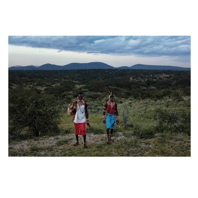 Two Samburu people in traditional dress stand in a grassy landscape with hills in the background under a cloudy sky, eye-level view.