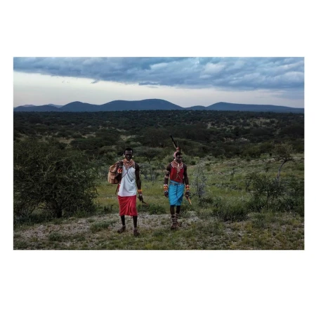 Two Samburu people in traditional dress stand in a grassy landscape with hills in the background under a cloudy sky, eye-level view.