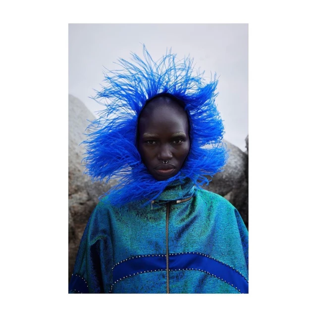 Striking fashion portrait of a dark-skinned adult woman wearing a bright blue feathered headdress and iridescent blue jacket outdoors.