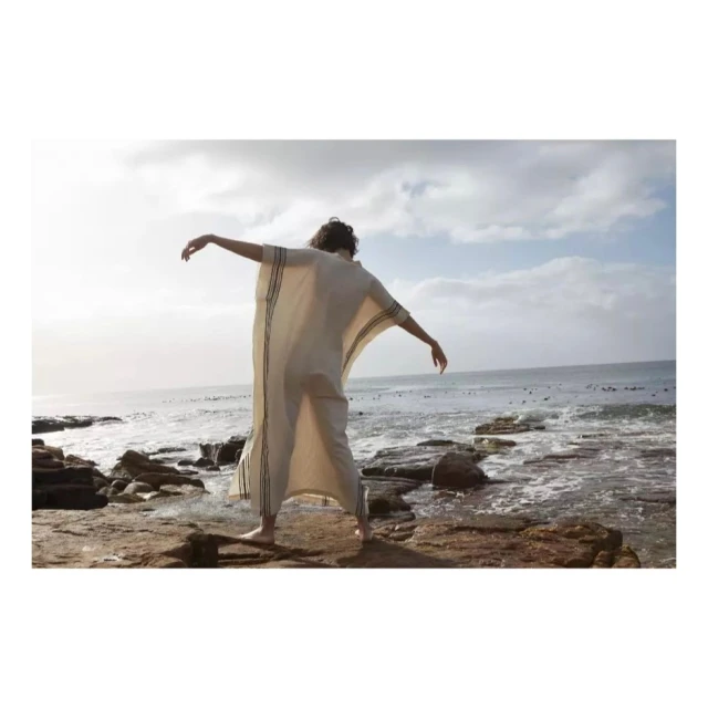 Back view of a person in a white caftan with arms outstretched on a rocky shore, ocean, and cloudy sky in the background.