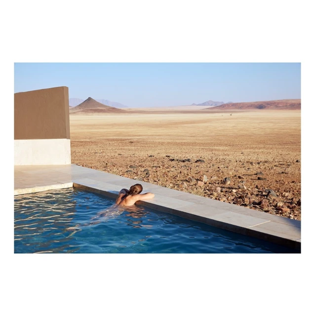 Woman relaxes in pool overlooking arid landscape. Brown earth, sparse vegetation, distant mountain under clear sky. Vacation, lifestyle.