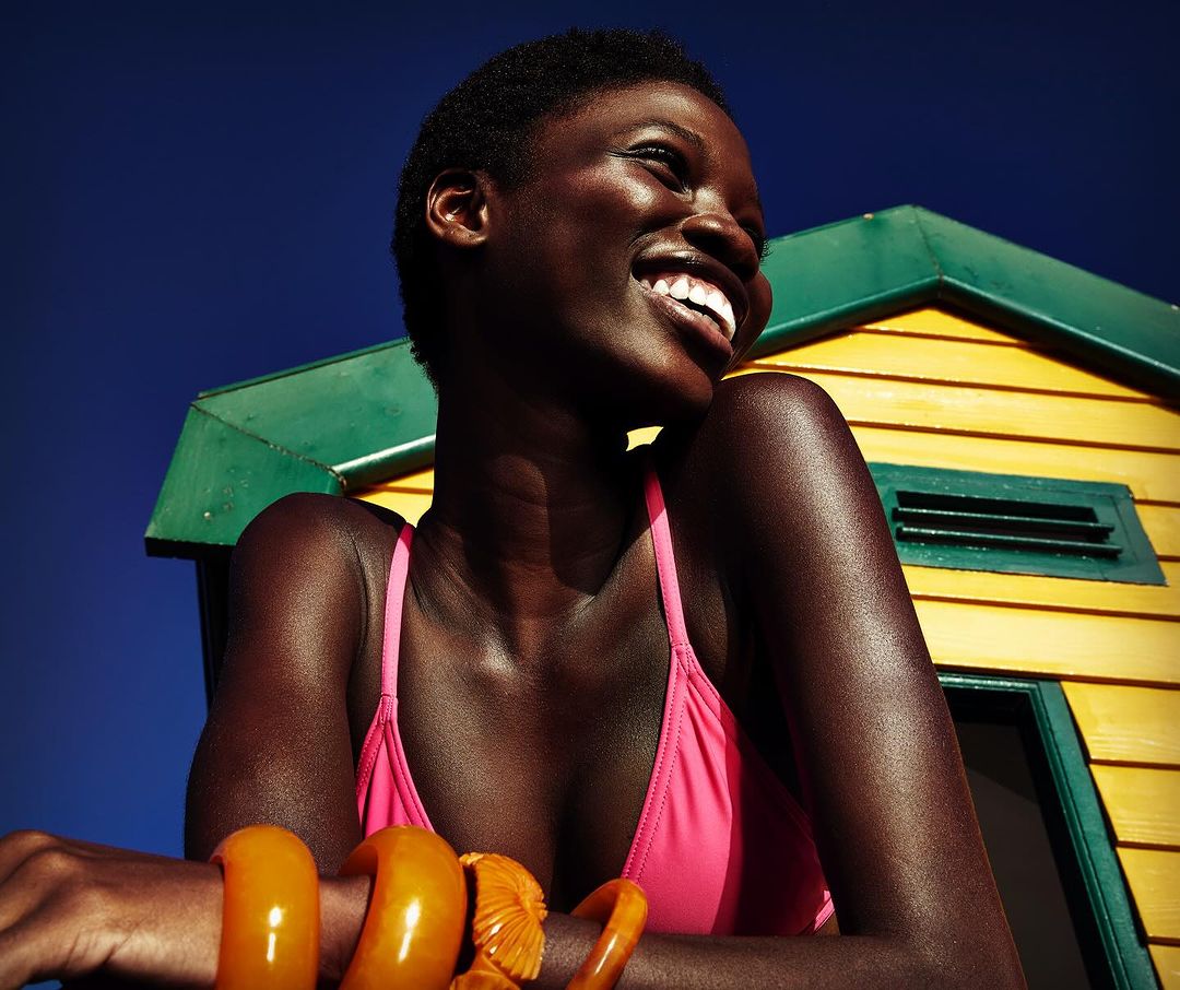 Smiling dark-skinned woman in pink top poses in front of yellow beach hut with orange bracelets in outdoor eye-level shot under blue sky.
