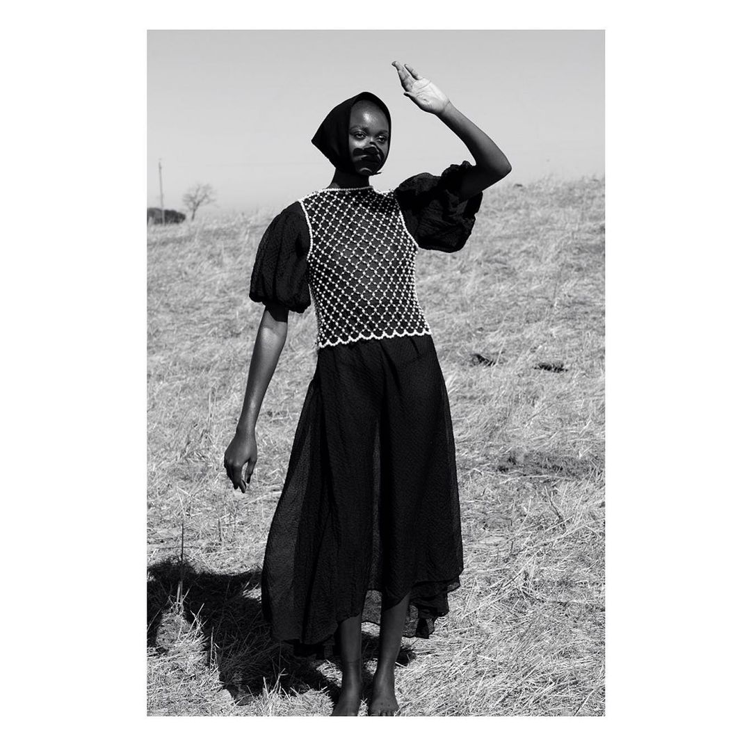 Black and white portrait of a Black female model shielding her eyes in an open field, wearing a patterned top and a long, flowing skirt.