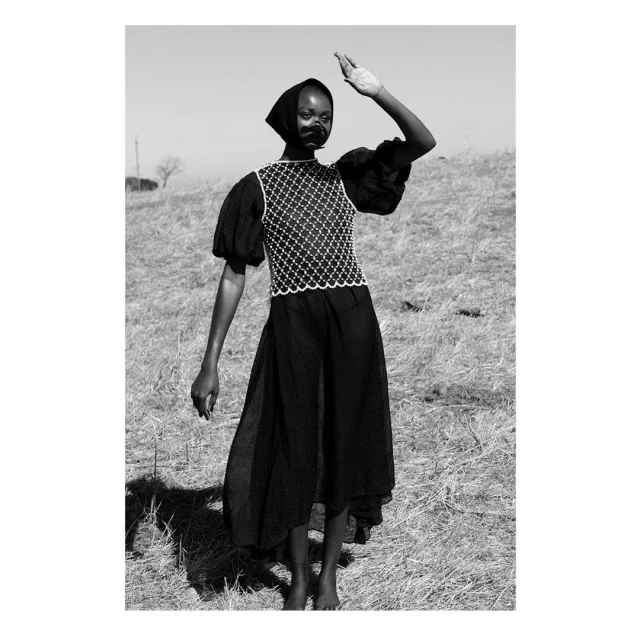 Black and white portrait of a Black female model shielding her eyes in an open field, wearing a patterned top and a long, flowing skirt.
