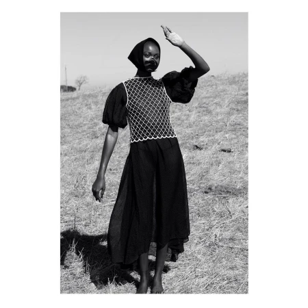 Black and white portrait of a Black female model shielding her eyes in an open field, wearing a patterned top and a long, flowing skirt.