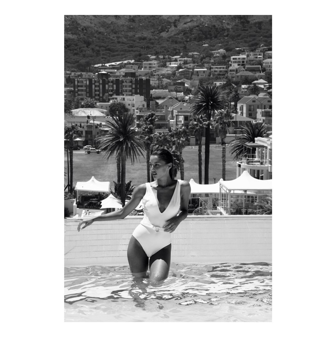 Woman in white swimsuit standing in an outdoor pool against a backdrop of coastal town and hills