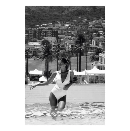 Woman in white swimsuit standing in an outdoor pool against a backdrop of coastal town and hills