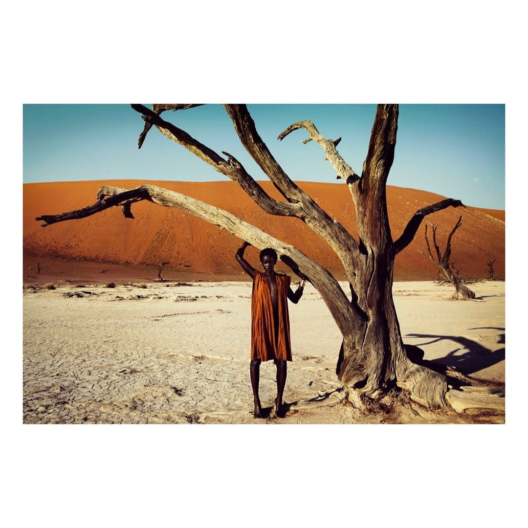 African woman in dress posing by a stark tree in a desert landscape with sand dunes under a clear blue sky, scenic portrait photography.
