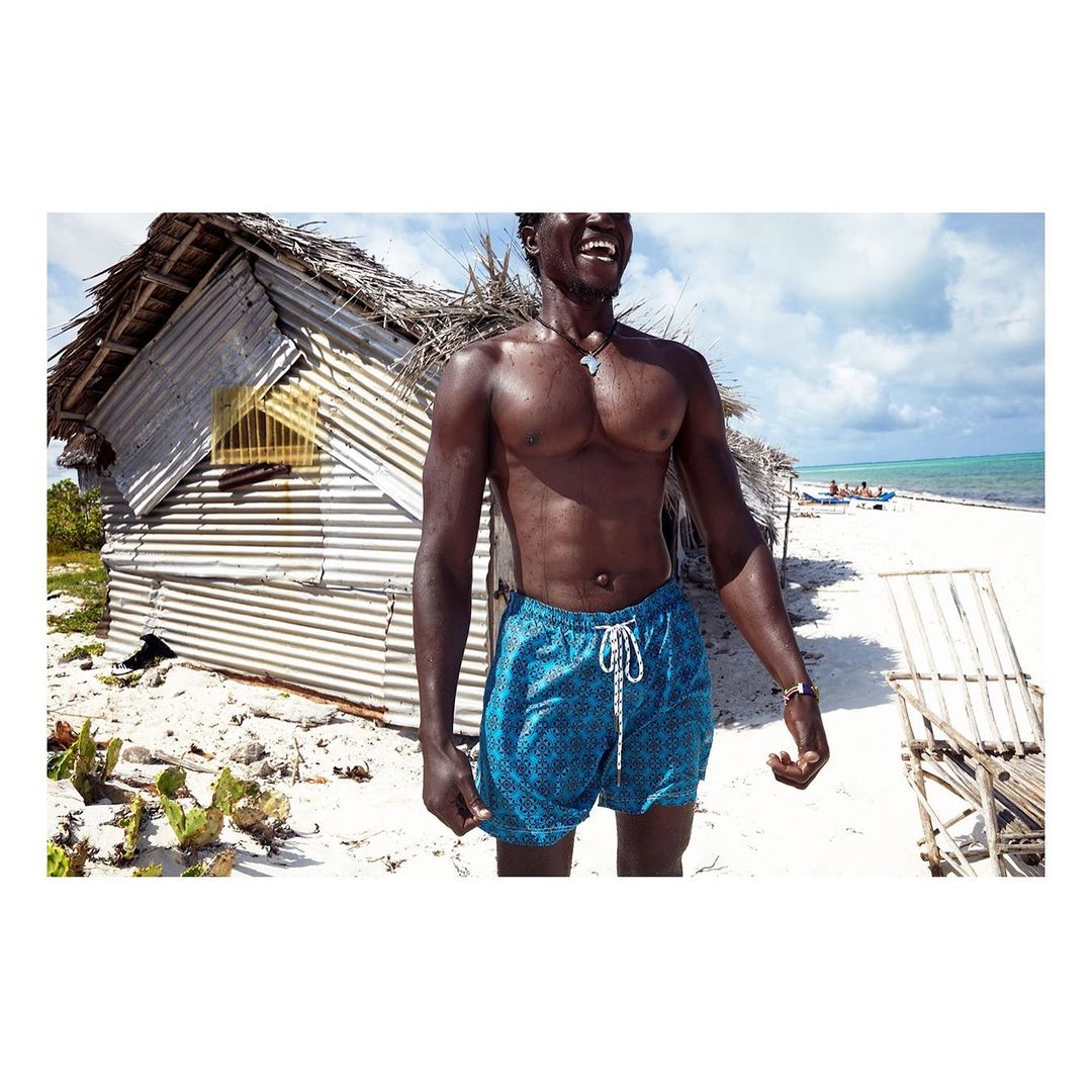 Smiling African man in blue swim trunks stands on a white sand beach near a weathered shack, with turquoise ocean and cloudy sky.