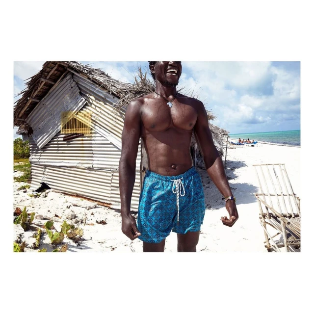 Smiling African man in blue swim trunks stands on a white sand beach near a weathered shack, with turquoise ocean and cloudy sky.