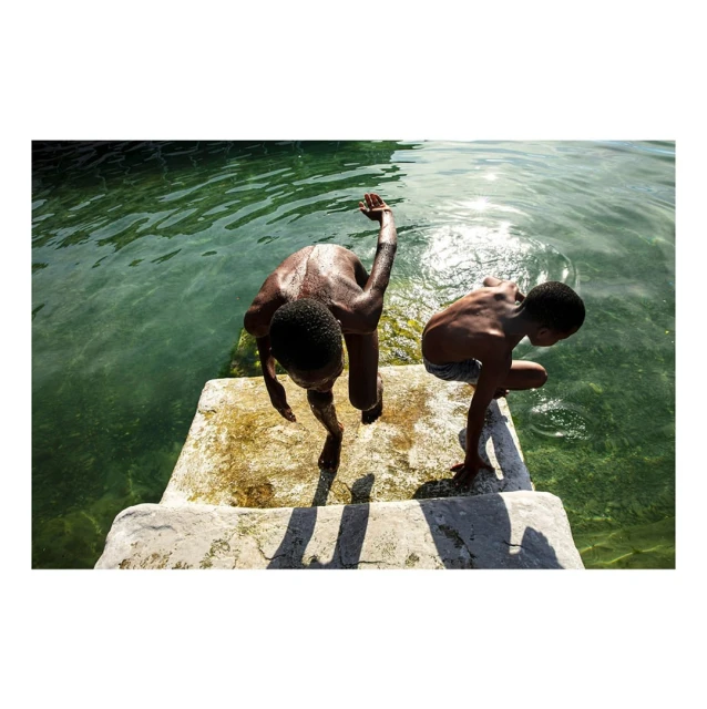 Two shirtless boys on a concrete platform prepare to jump into a clear body of water on a sunny day, captured from an overhead angle.